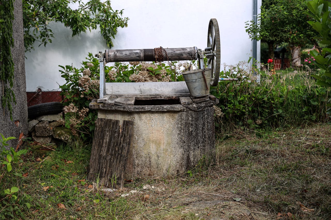 portfolio-03 A charming old stone well with a metal bucket set amidst lush garden greenery.