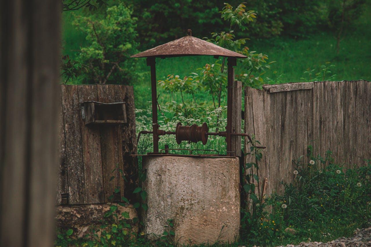 portfolio-04 A picturesque rustic well stands amidst lush greenery in a countryside garden, surrounded by a wooden fence.